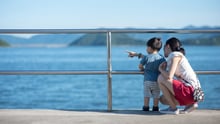 A mother holding a toddler points out towards the ocean.