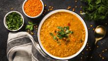 A bowl of lentil soup resting on a table, with two smaller bowls next to it filled with lentils and parsley.