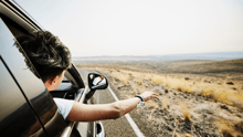 A person on a road trip in the car, sticking their arm and head out of the window with mountains in the distance.