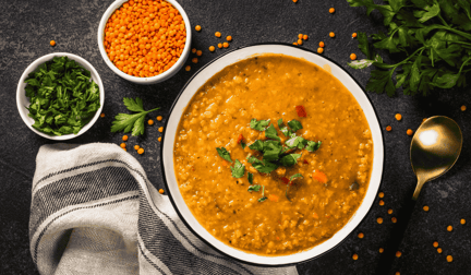 A bowl of lentil soup resting on a table, with two smaller bowls next to it filled with lentils and parsley.