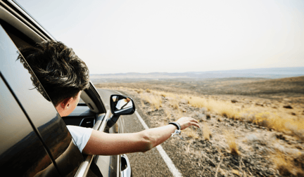 A person on a road trip in the car, sticking their arm and head out of the window with mountains in the distance.