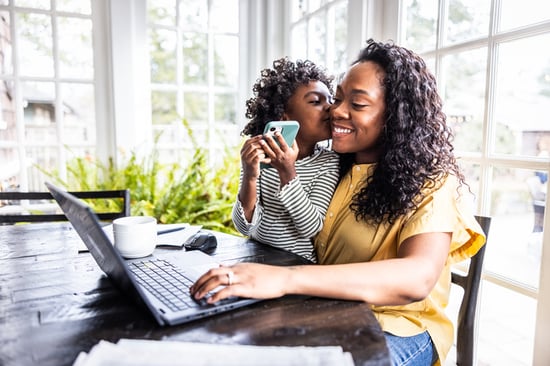 A mother smiles with a child on her lap with a laptop on a table.