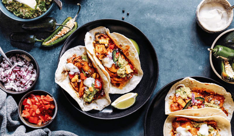 Plant-based tacos on a plate on a table with various topping surrounding them.