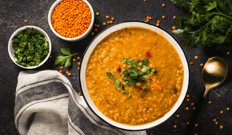 A bowl of lentil soup resting on a table, with two smaller bowls next to it filled with lentils and parsley.