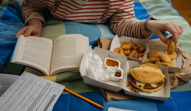 A college student eating fast food in bed while studying in their dorm.