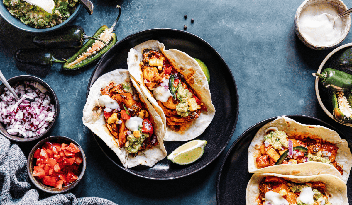 Plant-based tacos on a plate on a table with various topping surrounding them.