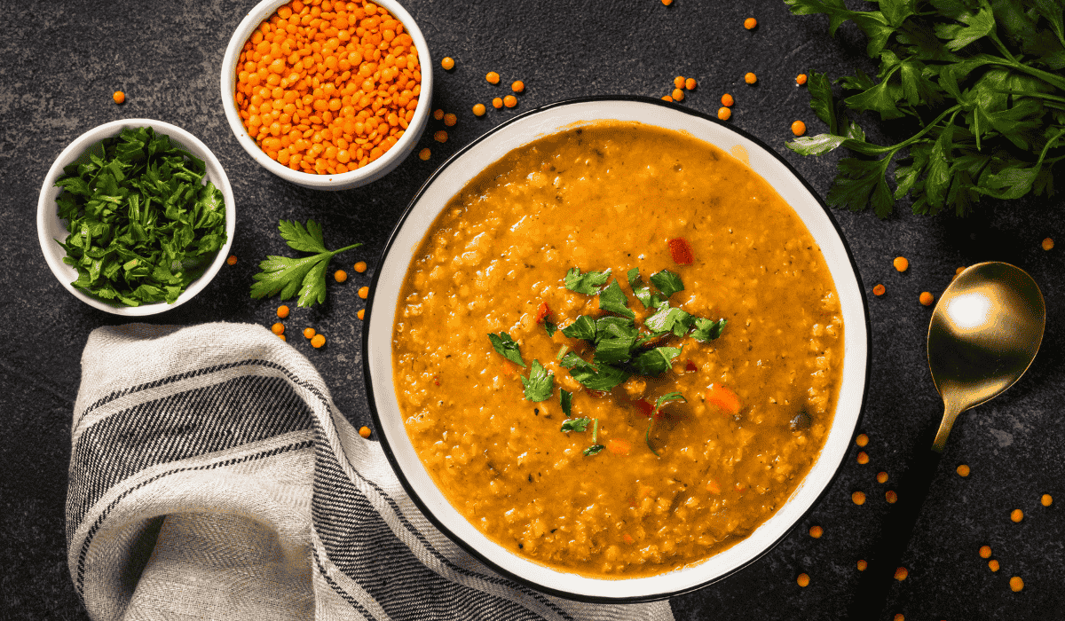 A bowl of lentil soup resting on a table, with two smaller bowls next to it filled with lentils and parsley.